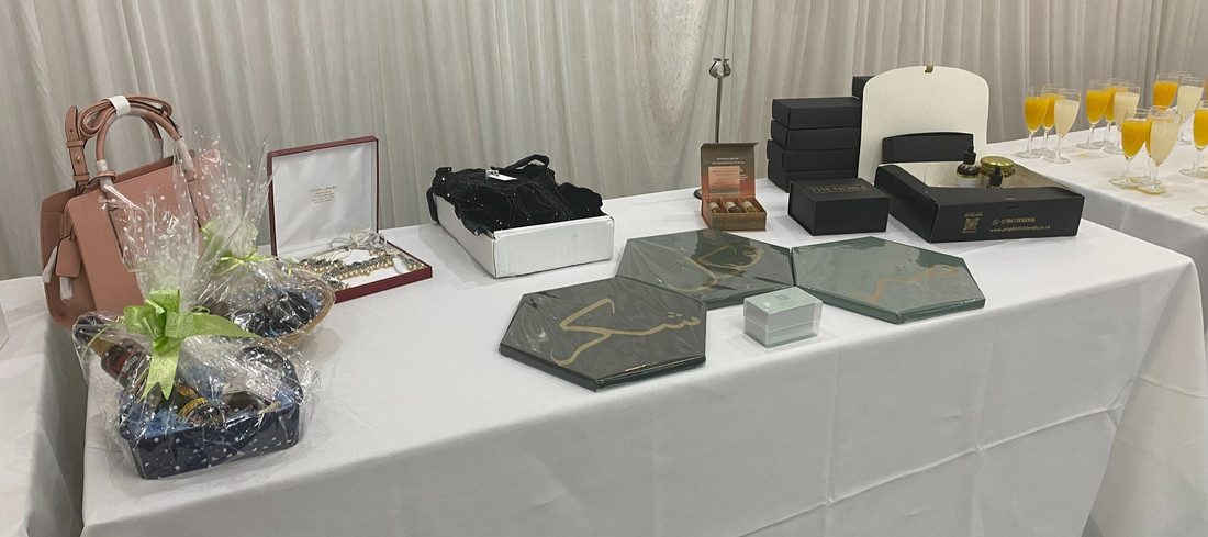 A gifts table with a pink handbag, cellophane-wrapped hampers, a jewellery box, slate coasters and glasses of orange juice.