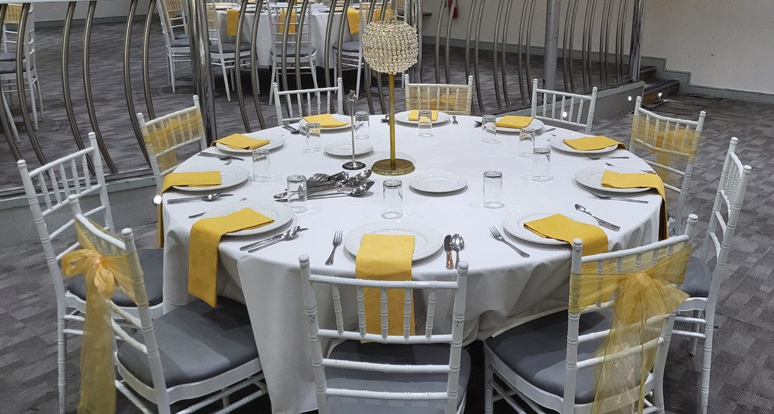 A round table laid for dinner with a white cloth, yellow napkins, a tall gold and crystal centrepiece, and white chairs tied with yellow sashes.