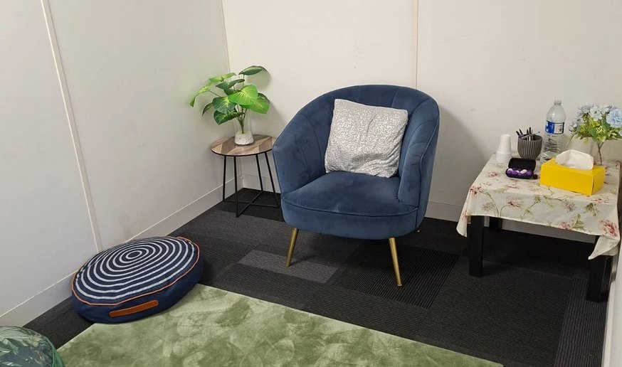 Our room: a blue velvet armchair with a cushion, a green rug with floor cushions, a plant on a side table, and a small table with tissues and flowers.
